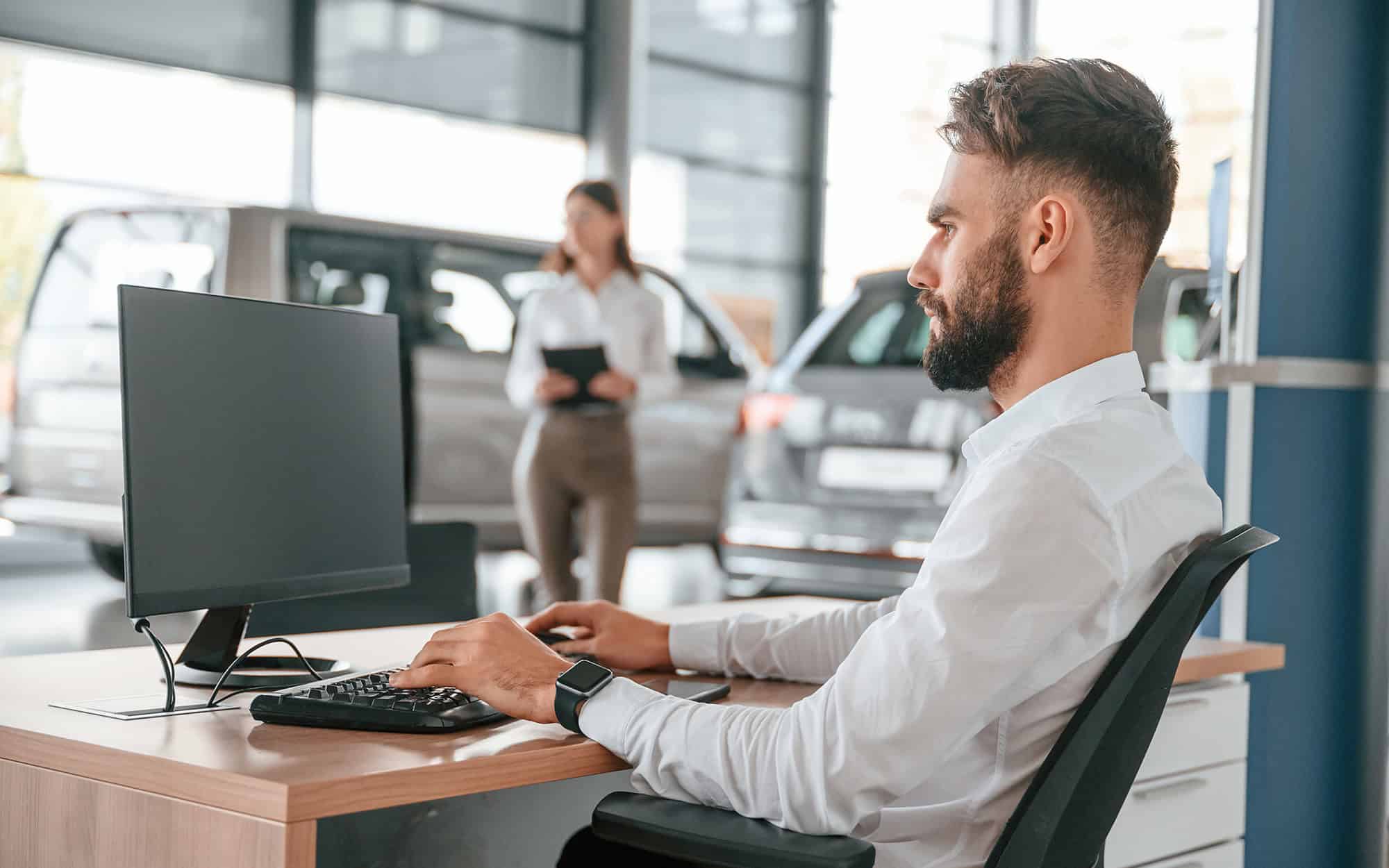 Side view of a man on his computer at a car dealership with woman in the background