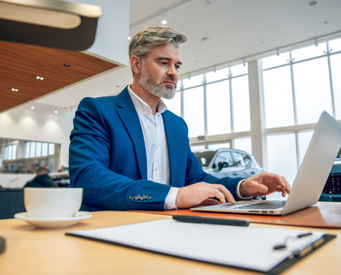 Bearded salesman in suit working on his laptop at new cars showroom