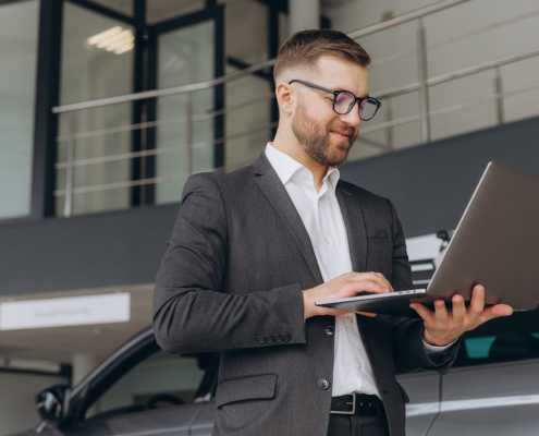 Modern bearded man in glasses and suit vehicle sales consultant using laptop inside car dealership